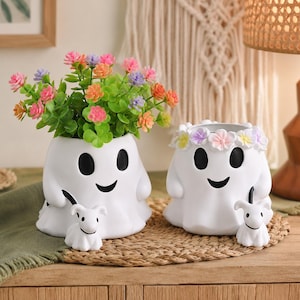 May include: Two white ghost-shaped planters with black eyes and mouths. One planter holds a bouquet of pink, orange, and purple flowers. Both ghosts are accompanied by a small white ghost dog figure. The second ghost wears a floral crown of pastel flowers.