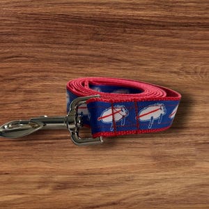 May include: A rolled-up dog leash with a blue and red pattern. The leash features a repeating design of white buffaloes with red accents on a blue background. A silver metal clasp is attached to the leash.