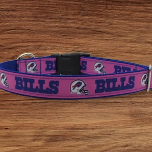 May include: Pink dog collar with blue and white Buffalo Bills logo and text. The collar has a silver buckle and a black plastic buckle.
