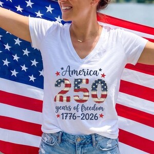 May include: A white V-neck t-shirt with the text "America 250 years of freedom 1776-2026" is worn in front of an American flag. The flag is red, white, and blue. The person is standing on a wooden dock near a lake.