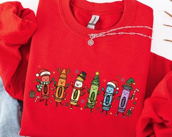 May include: A bright red crewneck sweatshirt featuring a graphic print of six colorful cartoon crayons dressed in festive holiday attire, including Santa hats, reindeer antlers, and scarves, with small candy canes and gift boxes.