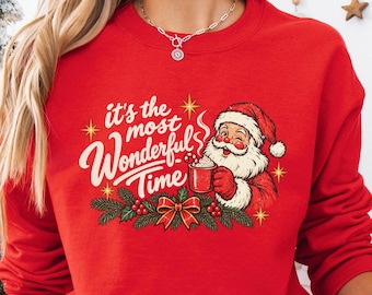 May include: A bright red long-sleeve sweatshirt featuring a graphic of Santa Claus holding a mug with the text "it's the most Wonderful Time" in white script, accented by green pine branches, red berries, and a red bow.