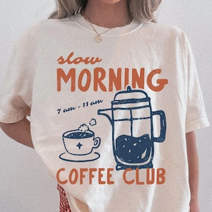May include: Ivory-colored t-shirt with a graphic design featuring the words "slow MORNING COFFEE CLUB" in orange and navy blue. The design includes a coffee cup, a French press, and the time "7 am - 11 am".