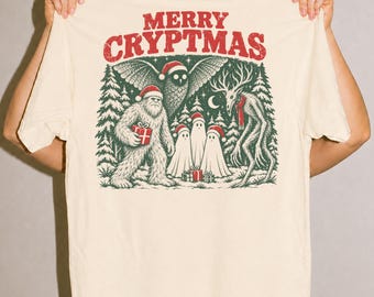May include: A cream-colored t-shirt featuring a graphic print with the text MERRY CRYPTMAS in red. The illustration depicts a Bigfoot, an owl, three ghosts, and a deer-like creature, all wearing Santa hats, standing in a forest.