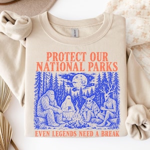 May include: A beige crewneck sweatshirt featuring a blue graphic print of a campfire scene with a sasquatch, a ghost, a moth creature, and a jackalope. The text reads PROTECT OUR NATIONAL PARKS and EVEN LEGENDS NEED A BREAK.