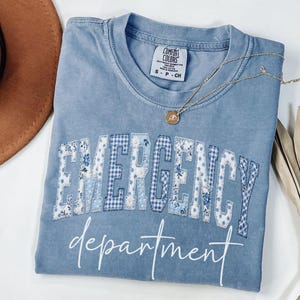 May include: A light blue t-shirt with the words "EMERGENCY department" in white script. The word "EMERGENCY" is in a patchwork design with floral and gingham patterns. A gold necklace with a pendant rests on the shirt.