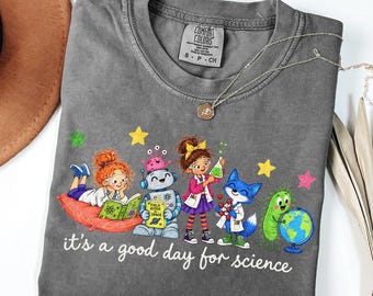 May include: A charcoal gray t-shirt featuring a graphic of two children, a robot, a fox, and a microbe engaged in scientific activities. The text reads, it's a good day for science. The shirt has a Comfort Colors label at the inner neckline.