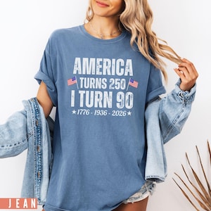 May include: Blue t-shirt with the text "AMERICA TURNS 250 I TURN 90" in white, with two small American flags. The shirt includes the years 1776-1936-2026. The model is wearing a denim jacket.