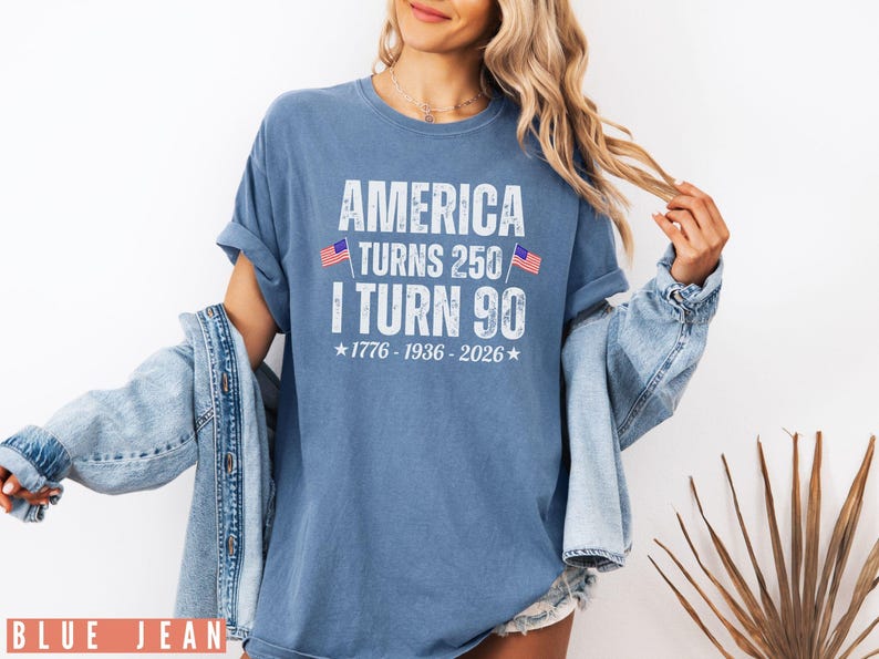 May include: Blue t-shirt with the text "AMERICA TURNS 250 I TURN 90" in white, with two small American flags. The shirt includes the years 1776-1936-2026. The model is wearing a denim jacket.