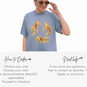 May include: A light blue t-shirt featuring a watercolor design of three golden goldfish with flowing fins and small gold accents. The shirt is a short-sleeved, crew neck style.
