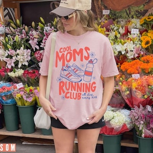 K&ouml;nnte beinhalten: Hellrosa T-Shirt mit dem Text "COOL MOMS RUNNING CLUB" in Rot und Blau, mit einer Grafik von Laufschuhen und einer Wasserflasche. Das Shirt wird vor einem Blumenstand getragen.