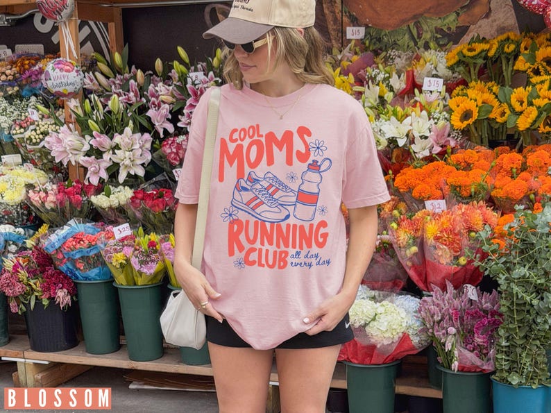 K&ouml;nnte beinhalten: Hellrosa T-Shirt mit dem Text "COOL MOMS RUNNING CLUB" in Rot und Blau, mit einer Grafik von Laufschuhen und einer Wasserflasche. Das Shirt wird vor einem Blumenstand getragen.