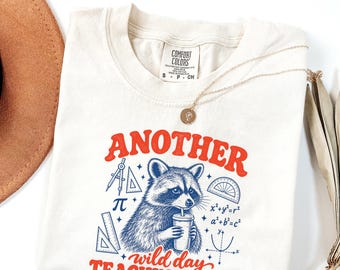 May include: A cream-colored t-shirt featuring a graphic of a raccoon holding a cup, surrounded by math symbols and geometry tools. The text reads "ANOTHER wild day TEACHING MATH" in red, with blue illustrations of a compass, protractor, and equations.