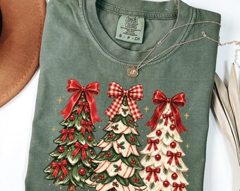 May include: An olive green crewneck t-shirt featuring a graphic of three Christmas trees decorated with red bows and ornaments. The center tree has a red and white checkered bow. The shirt has a Comfort Colors brand label in the neck.
