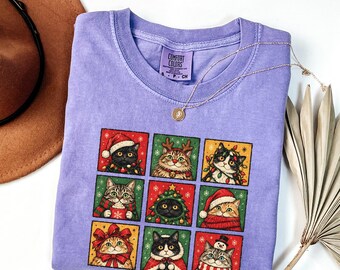 May include: A light purple crew neck t-shirt featuring a grid of nine illustrated cats in festive holiday attire, including Santa hats, scarves, and Christmas lights. The shirt has a Comfort Colors label at the neck and a short-sleeved design.