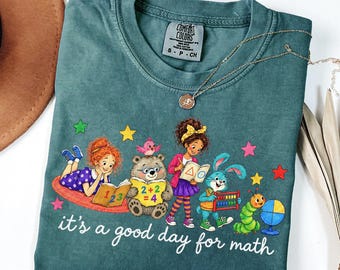 May include: A teal short-sleeve t-shirt featuring a graphic of children and animals studying math with the text "it's a good day for math". The shirt has a Comfort Colors label at the neck and is paired with a gold-toned chain necklace.