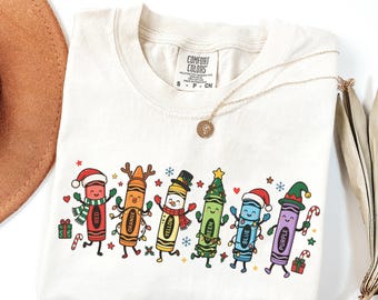 May include: A cream-colored t-shirt featuring a graphic of six anthropomorphic crayons labeled red, orange, yellow, green, blue, and purple. The crayons are decorated with holiday-themed accessories like Santa hats, reindeer antlers, and scarves, surrounded by small stars, snowflakes, and gift boxes.