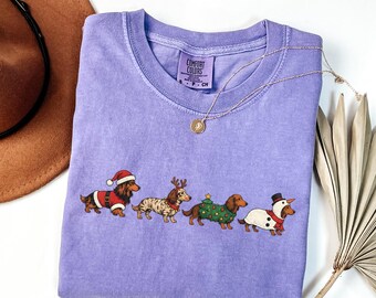 May include: A light purple short-sleeve t-shirt featuring a graphic of four dachshunds dressed in holiday costumes, including a Santa suit, reindeer antlers, a Christmas tree sweater, and a snowman outfit, with a Comfort Colors label.