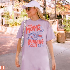 K&ouml;nnte beinhalten: Ein lila T-Shirt mit dem Text "COOL MOMS RUNNING CLUB" in Rot und Blau, mit Illustrationen von Turnschuhen und einer Wasserflasche. Das Shirt wird von einer Person getragen, die eine wei&szlig;e Kappe und Sonnenbrille tr&auml;gt.