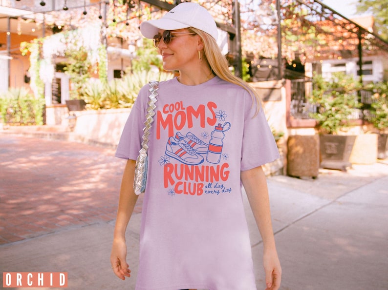 K&ouml;nnte beinhalten: Ein lila T-Shirt mit dem Text "COOL MOMS RUNNING CLUB" in Rot und Blau, mit Illustrationen von Turnschuhen und einer Wasserflasche. Das Shirt wird von einer Person getragen, die eine wei&szlig;e Kappe und Sonnenbrille tr&auml;gt.