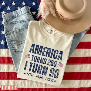 May include: An ivory-colored t-shirt with the text "AMERICA TURNS 250 I TURN 90" in navy blue, along with the years 1776-1936-2026 and small American flags. The shirt is paired with light-wash jeans and a tan hat, set against an American flag.