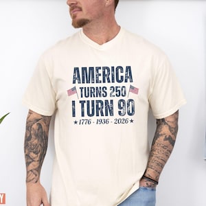 May include: Ivory-colored t-shirt with the text "AMERICA TURNS 250 / I TURN 90" in navy blue, flanked by small American flags. The shirt also includes the years "1776-1936-2026" below the text. The shirt is a v-neck style.