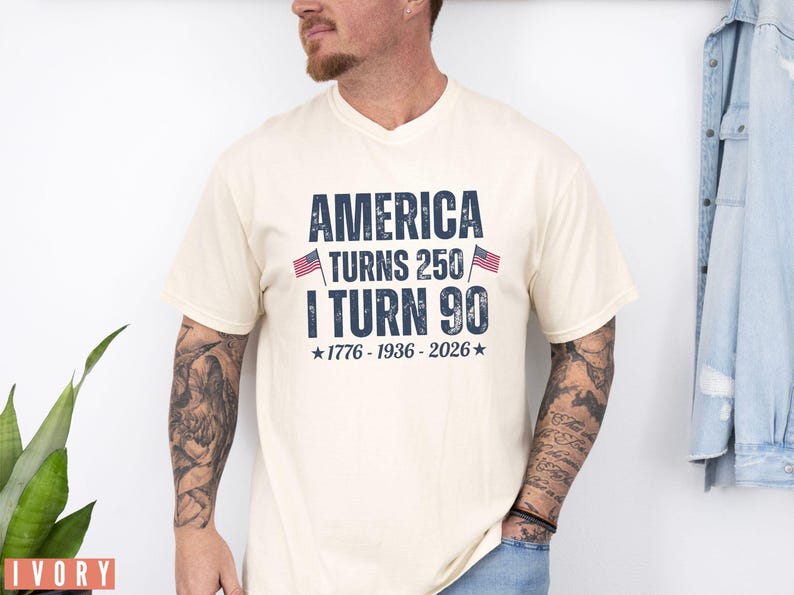 May include: Ivory-colored t-shirt with the text "AMERICA TURNS 250 / I TURN 90" in navy blue, flanked by small American flags. The shirt also includes the years "1776-1936-2026" below the text. The shirt is a v-neck style.