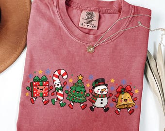 May include: A dusty rose t-shirt featuring a graphic print of five animated Christmas characters: a gift box, a candy cane, a pine tree, a snowman, and a bell. The shirt has a Comfort Colors brand label at the inner neckline.