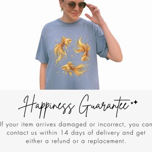 May include: A light blue t-shirt featuring a watercolor design of three golden goldfish. The fish have flowing fins and are surrounded by small gold dots. The shirt is a casual style.