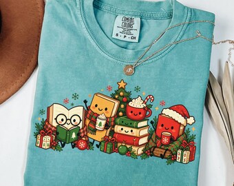 May include: A teal crewneck t-shirt with a Comfort Colors label featuring a graphic of anthropomorphic books, a Christmas tree, and a mug of cocoa. The illustration includes festive elements like wrapped gifts, snowflakes, and ornaments.