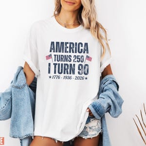 May include: White t-shirt with the text "AMERICA TURNS 250 I TURN 90" in navy blue. The shirt includes the years 1776-1936-2026 and small American flags. The shirt is worn with a denim jacket and shorts.
