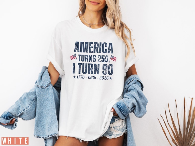 May include: White t-shirt with the text "AMERICA TURNS 250 I TURN 90" in navy blue. The shirt includes the years 1776-1936-2026 and small American flags. The shirt is worn with a denim jacket and shorts.