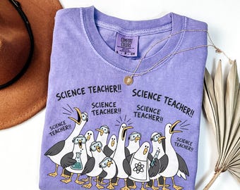 May include: A light purple t-shirt featuring a graphic of multiple seagulls wearing safety goggles and lab coats, with the text "SCIENCE TEACHER!!" repeated several times. The shirt has a Comfort Colors brand label on the inner neckline.