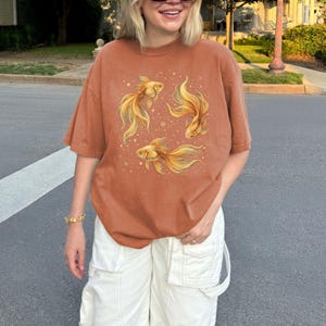 May include: Rust-colored t-shirt featuring a watercolor illustration of three golden fish with flowing fins and small stars. The shirt has a relaxed fit and short sleeves. The model is wearing white cargo pants.