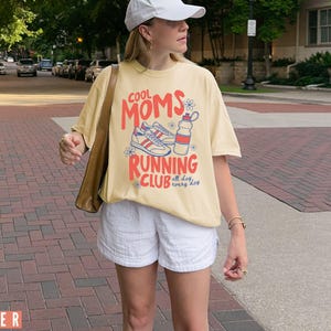 K&ouml;nnte beinhalten: Hellgelbes T-Shirt mit dem Aufdruck "COOL MOMS RUNNING CLUB" in Rot und Orange, zusammen mit einer Grafik von Turnschuhen und einer Wasserflasche. Das Shirt wird mit wei&szlig;en Shorts kombiniert. Der Text "all day, every day" ist ebenfalls vorhanden.
