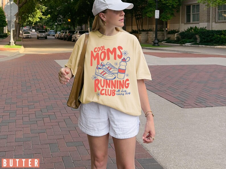 K&ouml;nnte beinhalten: Hellgelbes T-Shirt mit dem Aufdruck "COOL MOMS RUNNING CLUB" in Rot und Orange, zusammen mit einer Grafik von Turnschuhen und einer Wasserflasche. Das Shirt wird mit wei&szlig;en Shorts kombiniert. Der Text "all day, every day" ist ebenfalls vorhanden.