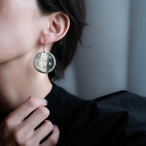 以下が含まれることがあります： 銀色の円形ドロップピアス。交差する線と星のような形が組み合わさった、繊細で複雑な幾何学模様の切り抜きが施されています。シンプルな金属製のフックから吊り下げられており、磨き上げられた金属の光沢と平らな円盤状のデザインが特徴です。