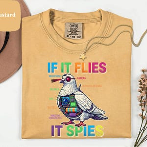 May include: Mustard-colored t-shirt with the text "IF IT FLIES IT SPIES" and a graphic of a pigeon with tech components. The shirt has a crew neck and a Comfort Colors tag.