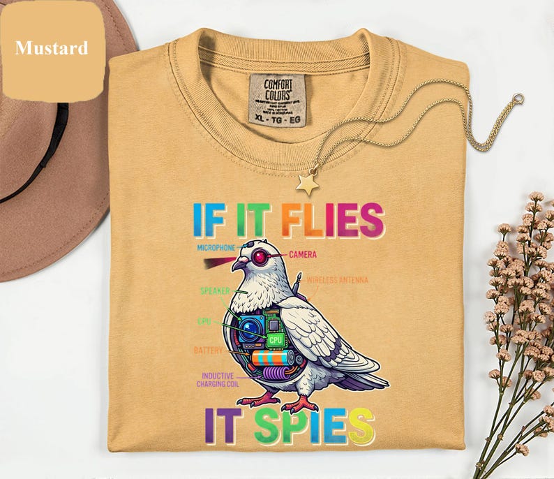 May include: Mustard-colored t-shirt with the text "IF IT FLIES IT SPIES" and a graphic of a pigeon with tech components. The shirt has a crew neck and a Comfort Colors tag.