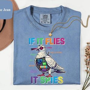 May include: A light blue t-shirt with the text "IF IT FLIES IT SPIES" and a graphic of a pigeon with tech components. The shirt has a crew neck and short sleeves. A gold necklace with a star pendant is on the shirt.