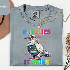 May include: A light blue Comfort Colors t-shirt with the text "IF IT FLIES IT SPIES" and a detailed illustration of a pigeon with spy gadgets. The shirt has a crew neck and short sleeves. A gold necklace with a star pendant is draped on the shirt.