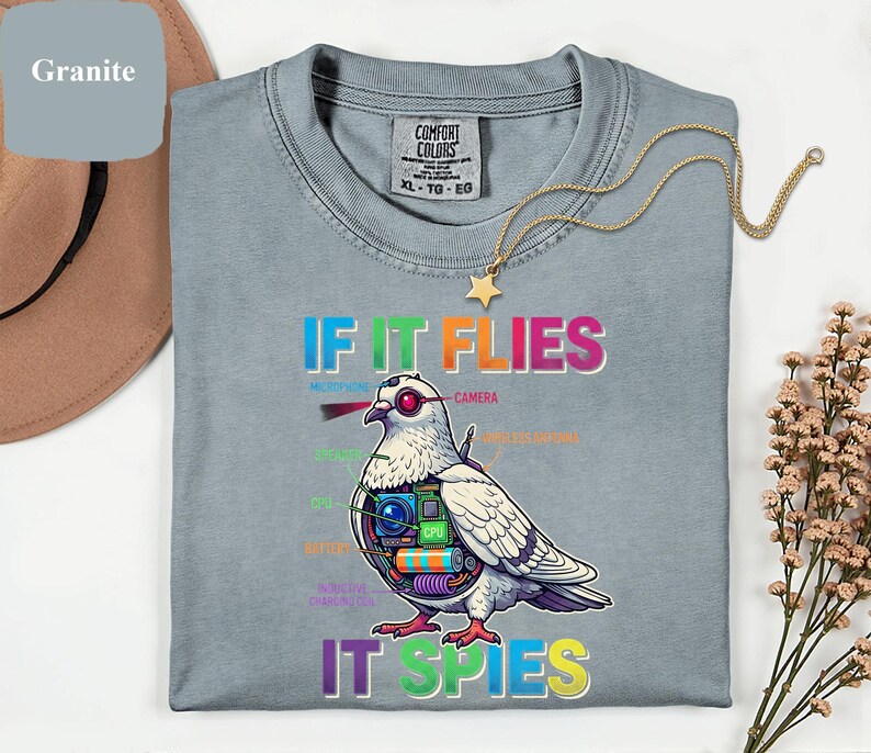 May include: A light blue Comfort Colors t-shirt with the text "IF IT FLIES IT SPIES" and a detailed illustration of a pigeon with spy gadgets. The shirt has a crew neck and short sleeves. A gold necklace with a star pendant is draped on the shirt.