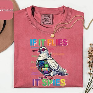 May include: A watermelon-colored t-shirt with the text "IF IT FLIES IT SPIES" and a graphic of a pigeon with tech components. The shirt has a crew neck and a gold star pendant necklace.