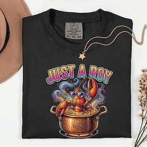 May include: Black t-shirt with a graphic of a lobster in a pot, wearing sunglasses, and the text "JUST A BOY". A gold star pendant necklace is draped over the shirt. The t-shirt is a Comfort Colors brand.