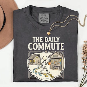 May include: Dark gray t-shirt with the text "THE DAILY COMMUTE" and a graphic of a person walking towards a workshop. The graphic includes a house, tools, and the text "EST. 2024". A gold star pendant necklace is draped across the shirt.