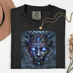 May include: Black crew neck t-shirt with a graphic of a stylized face with blue glowing eyes and the text "SHADOW ENTITY". A gold necklace with a star pendant rests on the shirt. The shirt has a Comfort Colors tag.