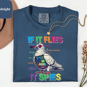 May include: Midnight blue t-shirt with a graphic of a pigeon with tech components and the text "IF IT FLIES IT SPIES". The shirt also has the Comfort Colors brand tag. A gold necklace with a star pendant is also visible.