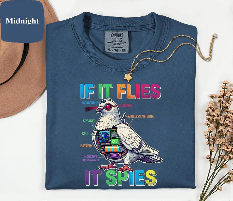 May include: Midnight blue t-shirt with a graphic of a pigeon with tech components and the text "IF IT FLIES IT SPIES". The shirt also has the Comfort Colors brand tag. A gold necklace with a star pendant is also visible.