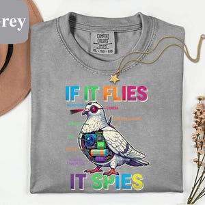 May include: A grey Comfort Colors XL t-shirt with the text "IF IT FLIES IT SPIES" and a graphic of a pigeon with tech components. A gold necklace with a star pendant is draped on the shirt.