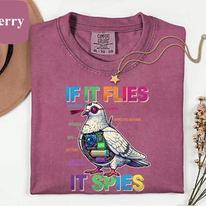 May include: Berry-colored t-shirt with the text "IF IT FLIES IT SPIES" and a graphic of a pigeon with tech components. The shirt has a crew neck and short sleeves. A gold necklace with a star pendant is on the shirt.